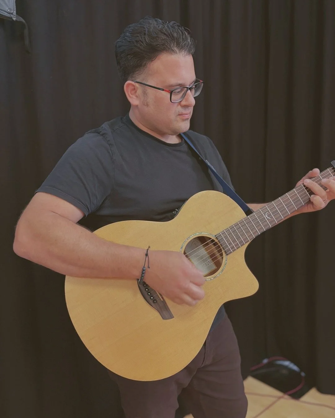 A man with glasses wearing a black t-shirt playing an acoustic guitar with a dark background.