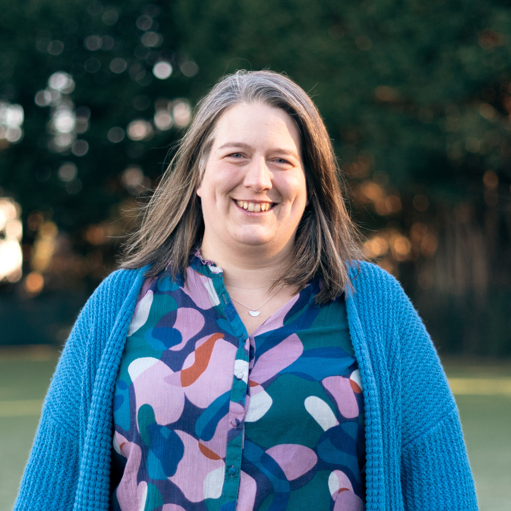 A woman with shoulder-length brown hair, wearing a blue textured sweater and a white chunky necklace, smiling at the camera against a black background.