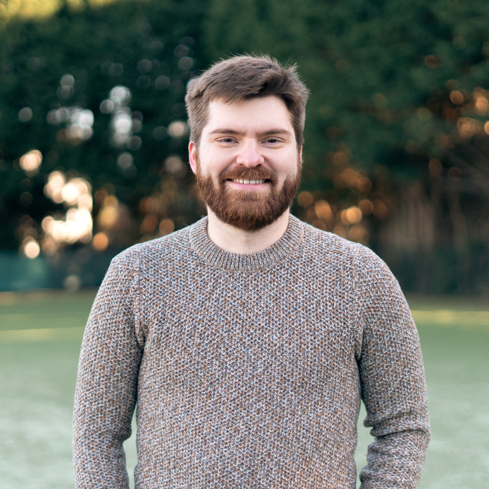 A smiling man and woman standing close together, with the woman wrapping her arm around the man's shoulders, against a dark background.