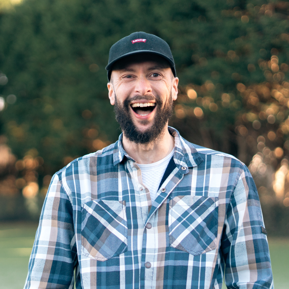 A smiling young woman with long brown hair and a young man with a beard and a leather jacket standing together against a black background.