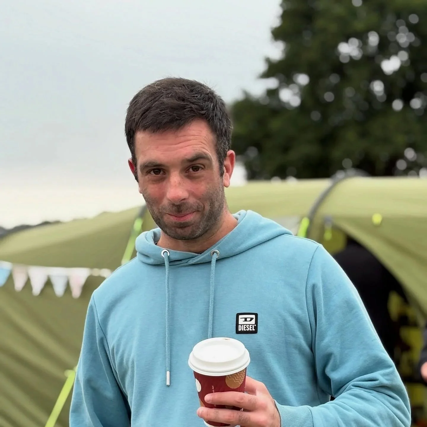 A man with dark hair and beard wearing a light blue hoodie holding a coffee cup outdoors, with tents and trees in the background.