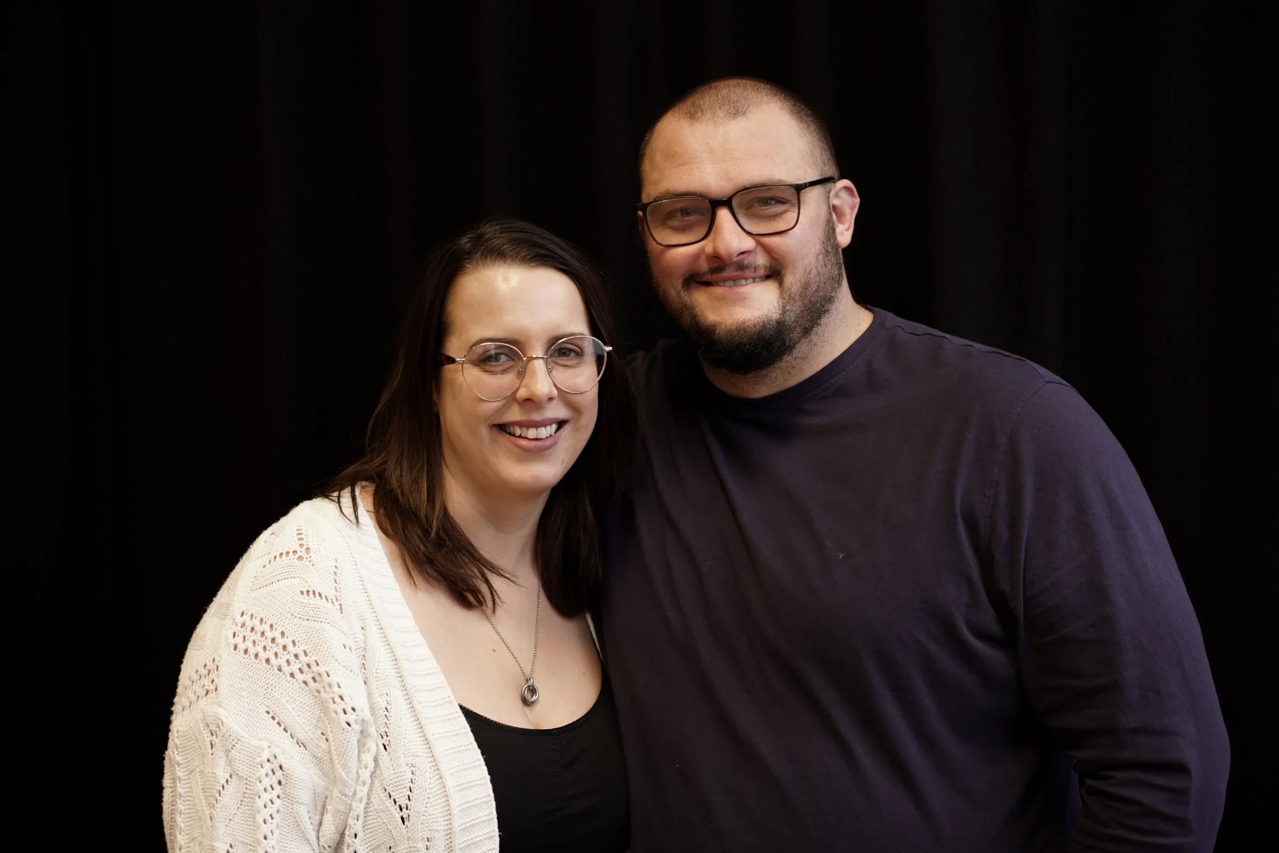 A smiling woman with shoulder-length brown hair and glasses, wearing a white knitted cardigan and a black top, standing next to a smiling man with short hair, beard, glasses, and a dark shirt, against a black background.