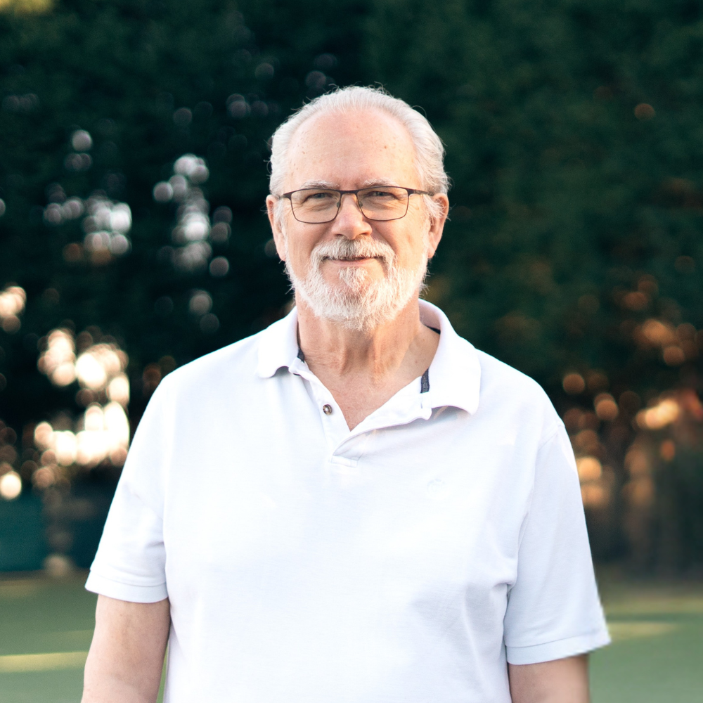 Older man with white hair, beard, and glasses smiling outdoors at sunset, wearing a white polo shirt.