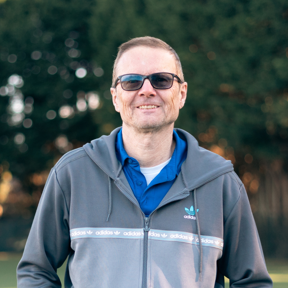 A man with glasses and a beard, smiling, wearing a light gray polo shirt with black and white accents, standing against a black background.
