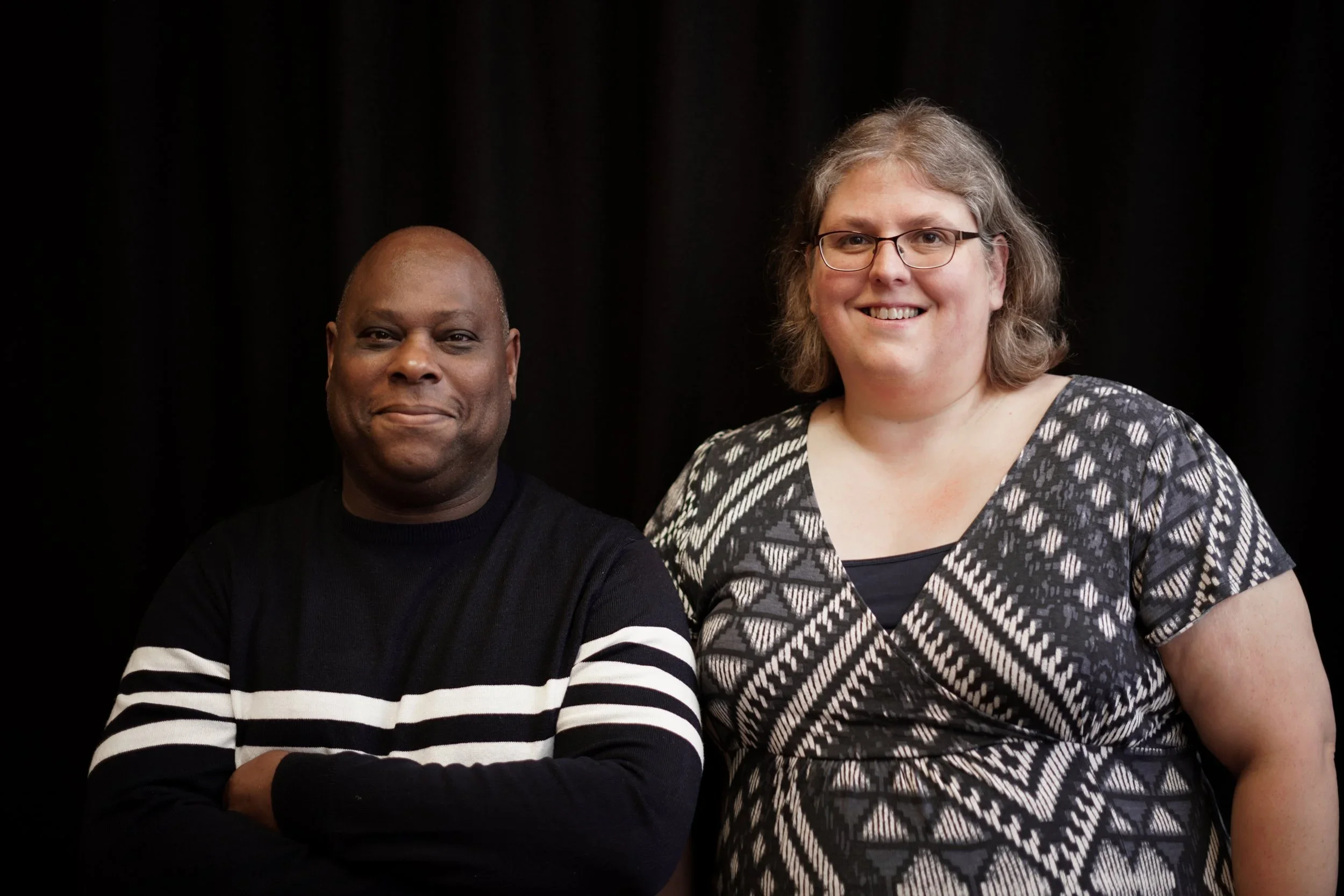 A man and a women standing side by side against a dark background, smiling at the camera.