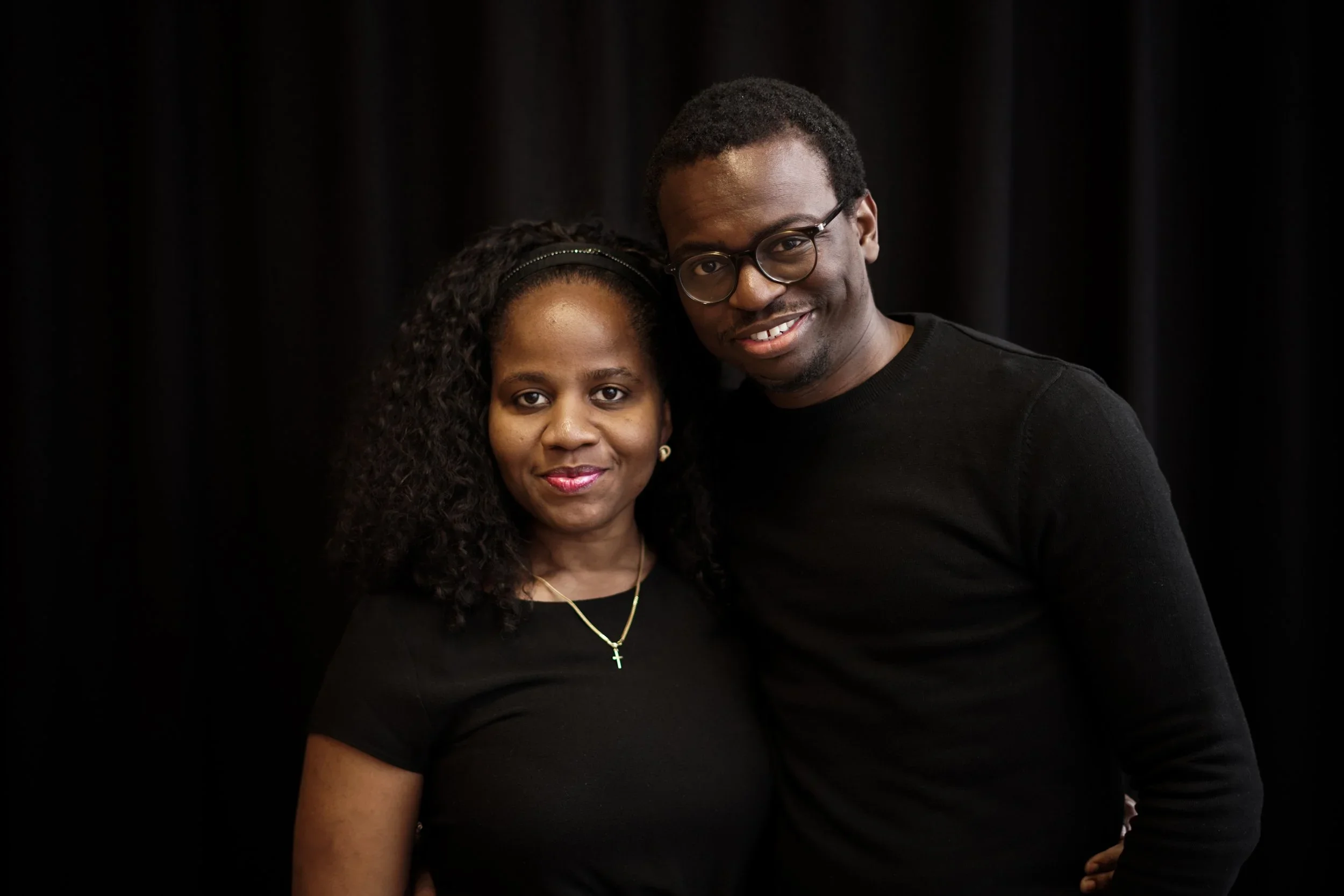 A man and woman standing close together in front of a black background, both wearing black shirts. The woman has curly hair, a necklace with a cross, and red lipstick. The man has short hair, glasses, and is smiling.