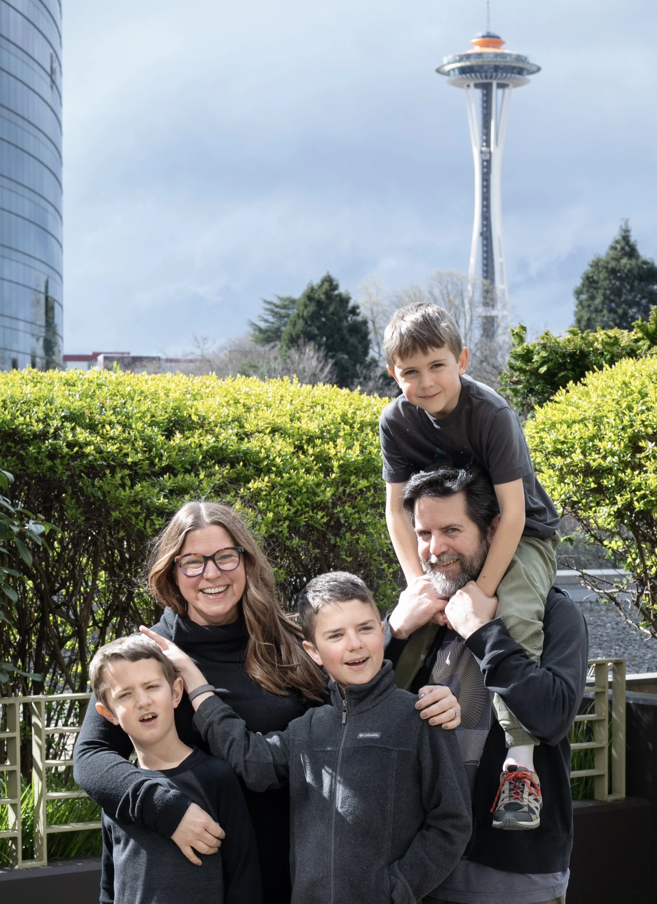 A family of five outdoors, with the Seattle Space Needle in the background. The family includes a woman with glasses, a man with a beard, and three boys. The woman and two boys are smiling and standing close together, while the man is carrying one of the boys on his shoulders. The setting appears to be a park or garden on a partly cloudy day.