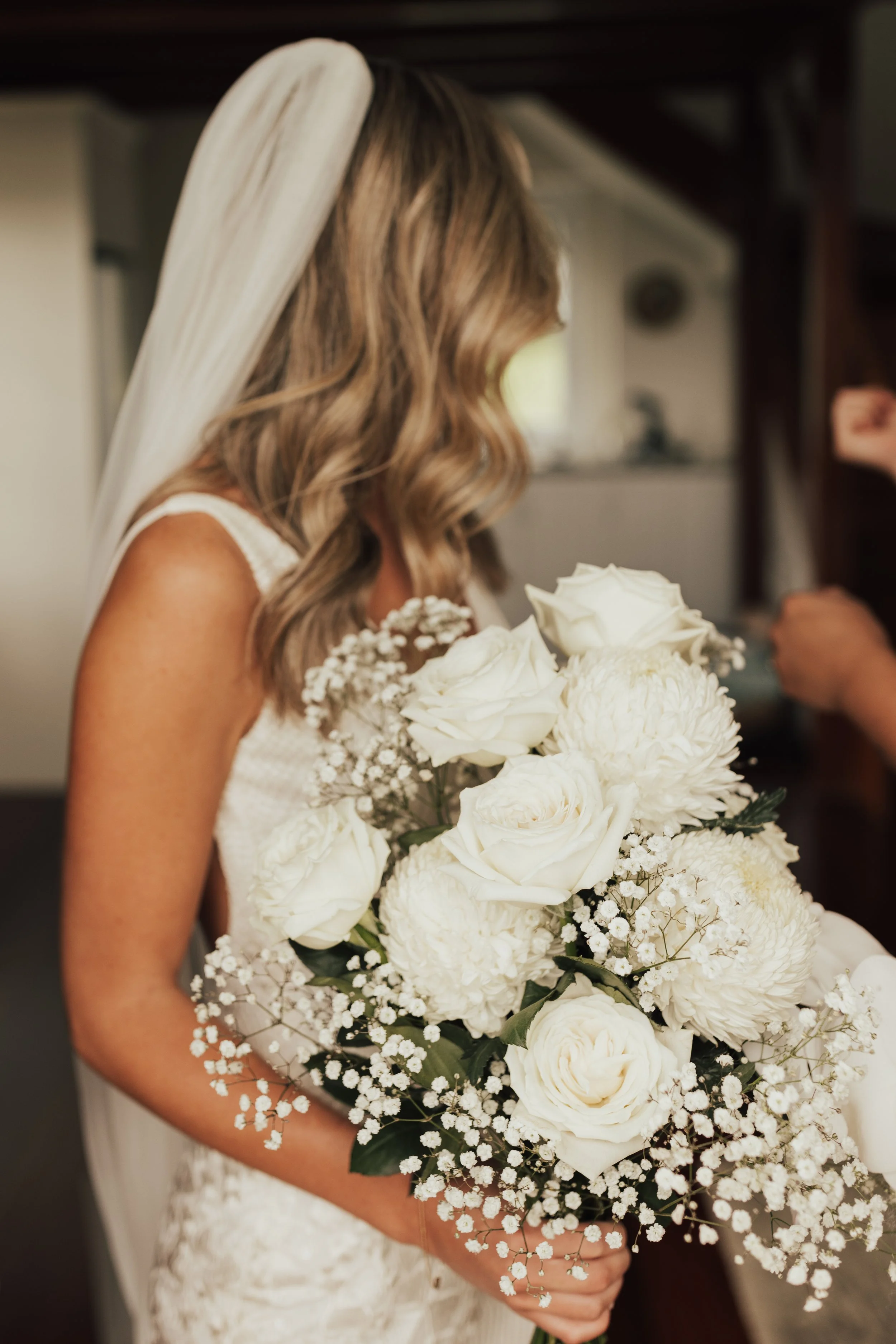 A bride holding a large bouquet of white roses, dahlias, baby's breath, and greenery, wearing a lace wedding dress and a veil, with her face slightly turned away.