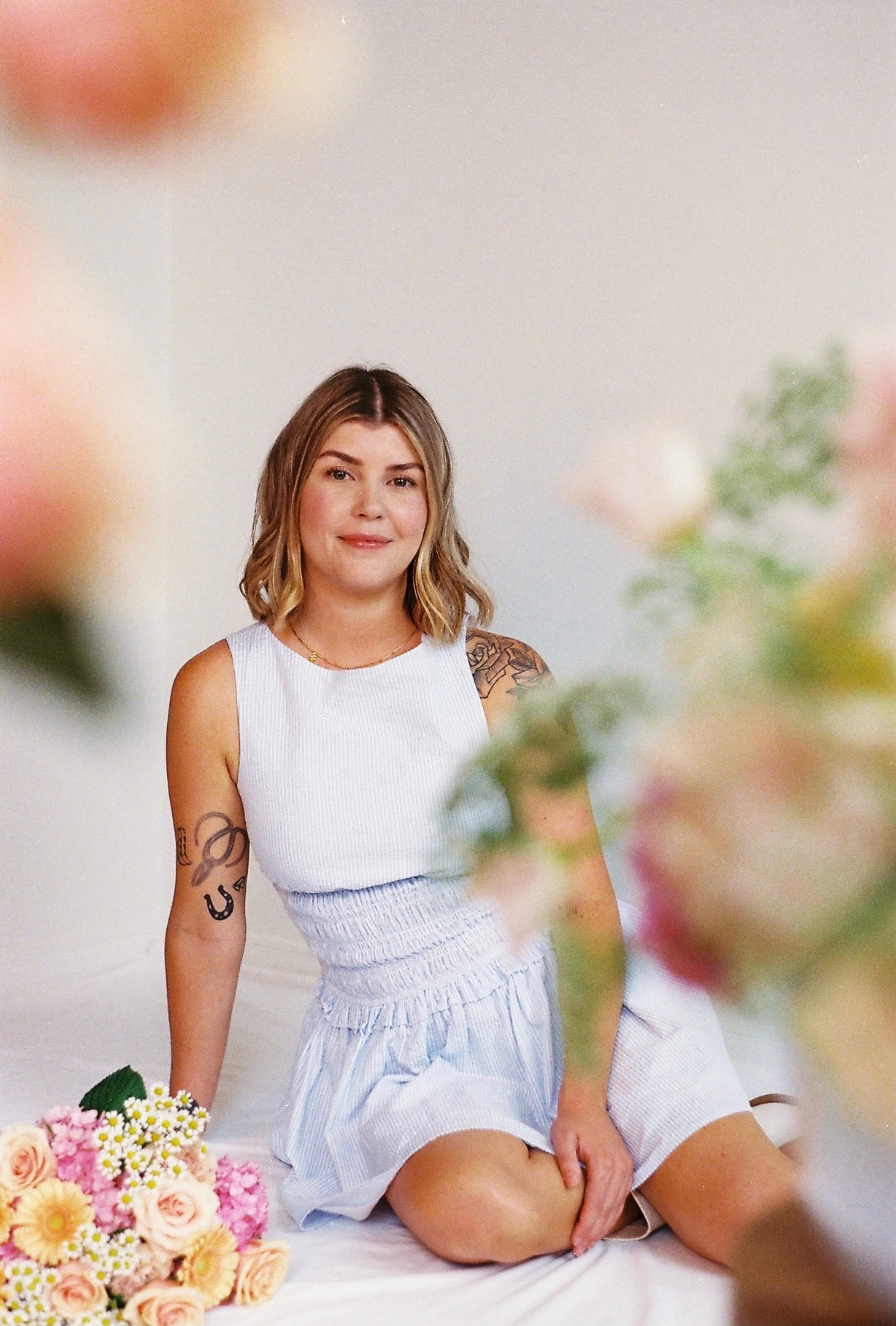 A young woman with shoulder-length wavy hair, wearing a blue sleeveless dress, sitting with a bouquet of pink and cream flowers nearby, and blurred flowers in the foreground.