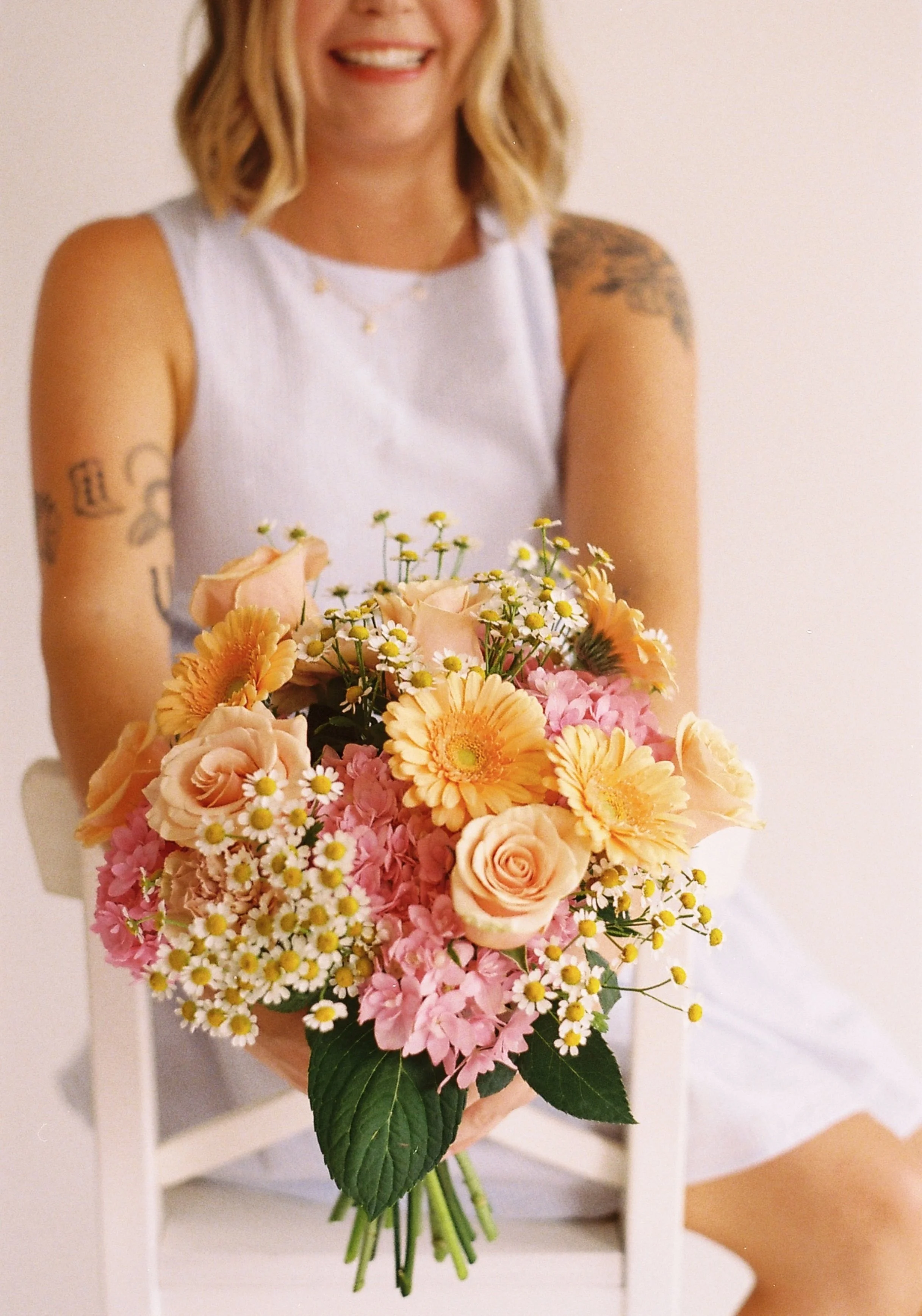 A woman sitting on a white chair holding a bouquet of pink, peach, and yellow flowers with a white background.