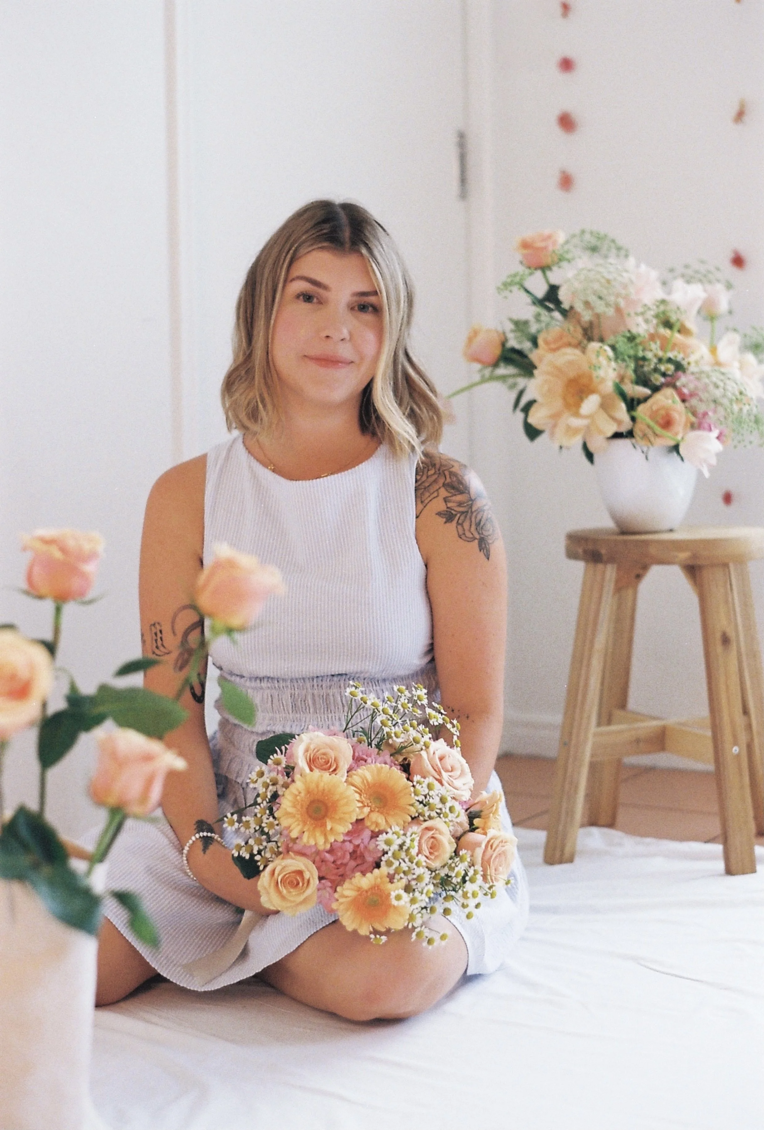 A young woman with shoulder-length wavy hair, wearing a white sleeveless top, holding a bouquet of peach and cream-colored flowers, sitting on a white bed with a background of white walls and a large floral arrangement on a wooden stand.