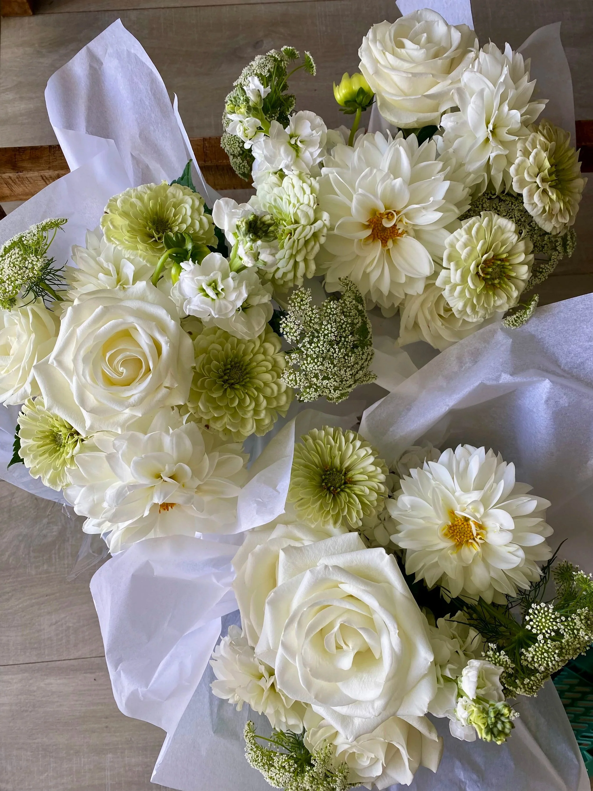 A bouquet of white flowers including roses, dahlias, and other blooms, wrapped in white paper, placed on a wooden surface.
