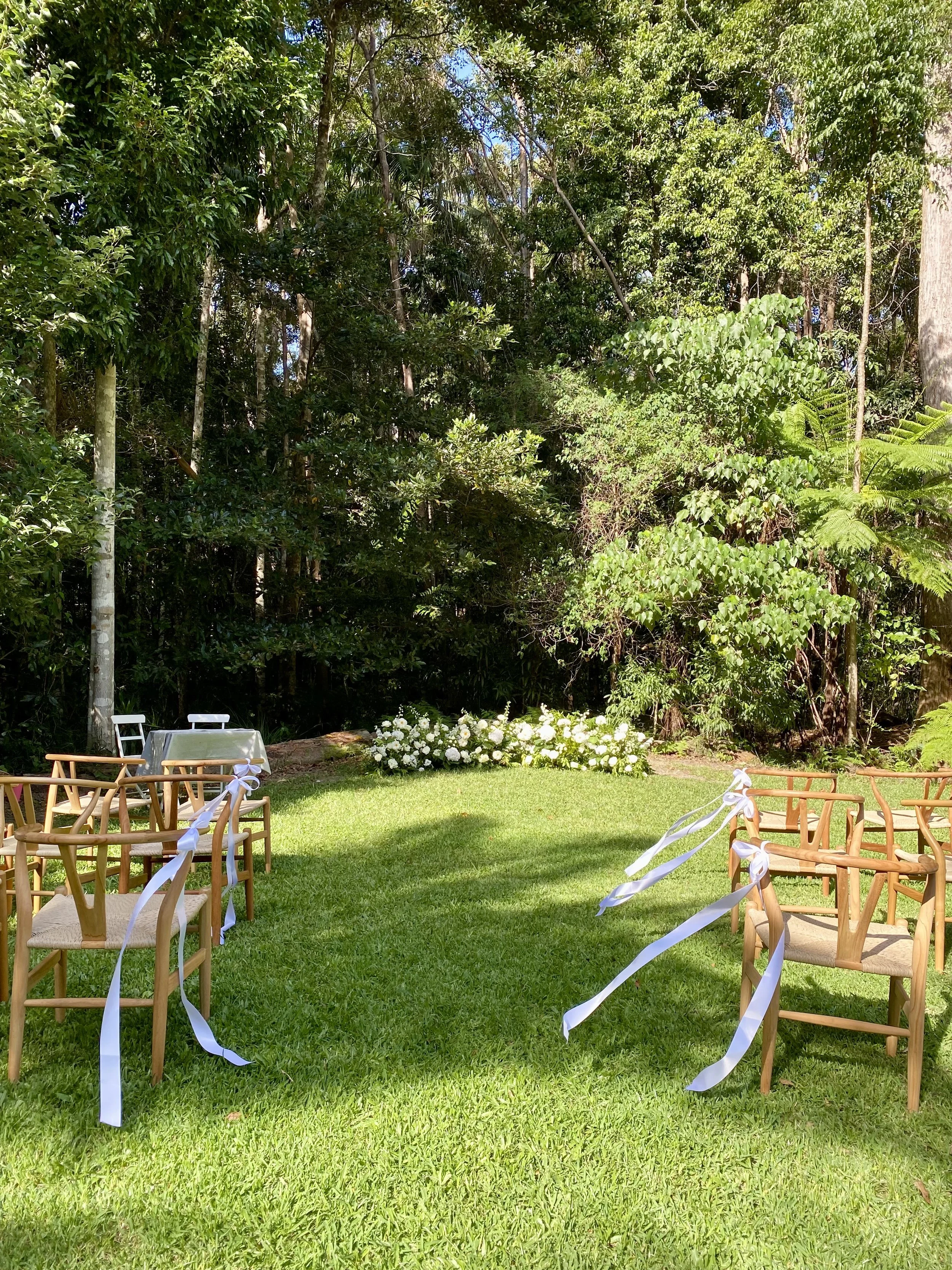 An outdoor wedding setup in a lush green garden with wooden chairs adorned with white ribbons, a white table, and a floral arrangement of white flowers in front of a backdrop of dense trees and foliage.