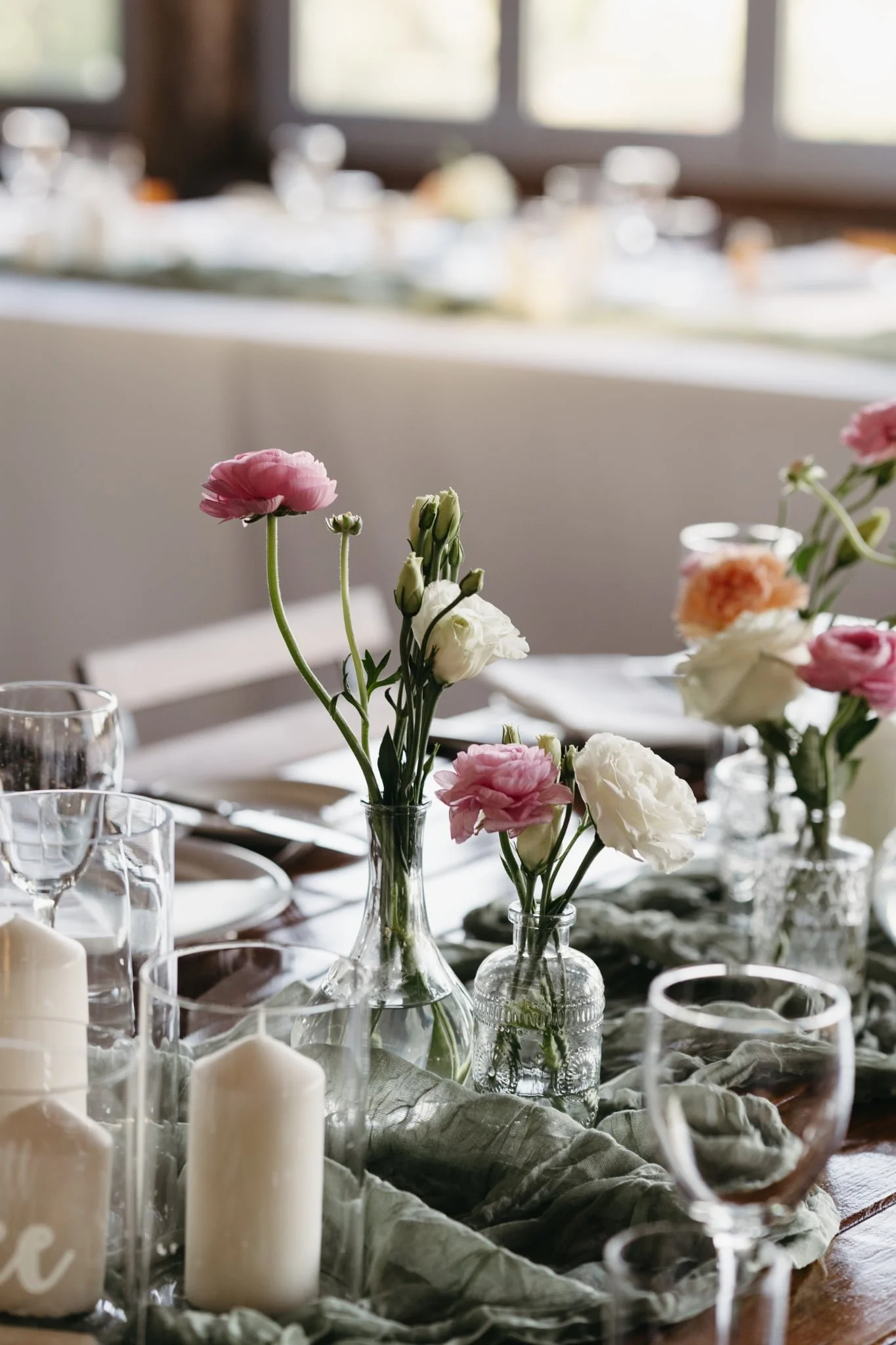 Elegant dining table decoration with glass vases holding pink and white flowers, candles in glass holders, and wine glasses on a green cloth table runner.