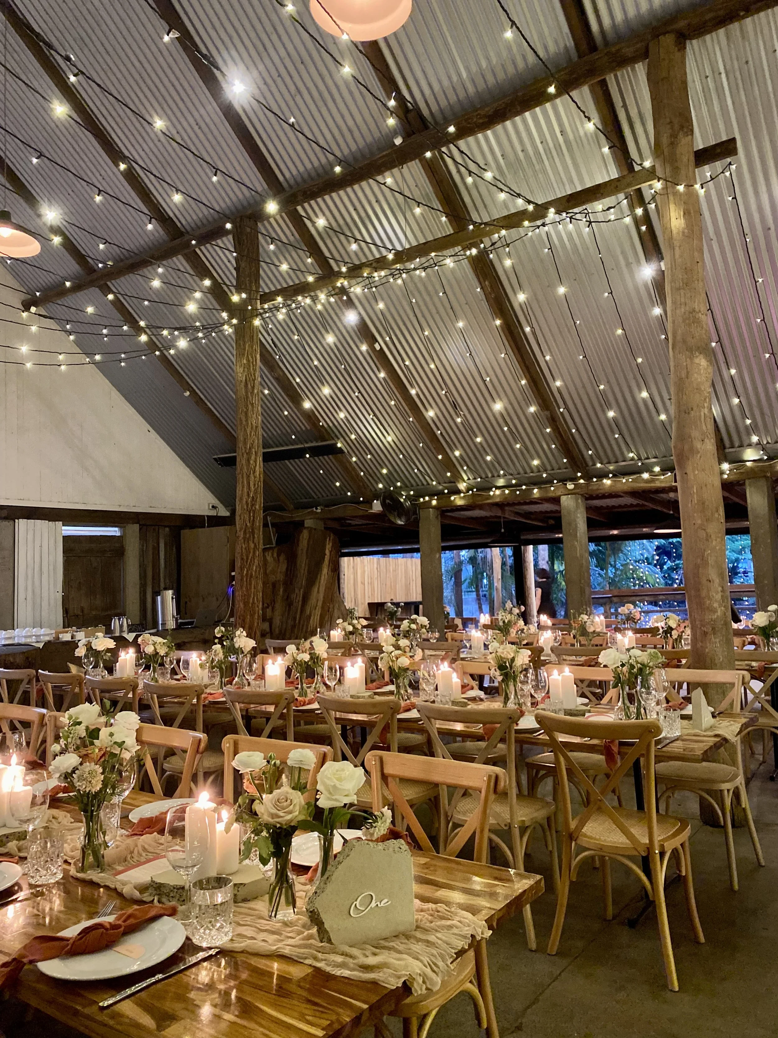 Decorated rustic wedding reception inside a barn with string lights, candles, floral centerpieces, and wooden tables and chairs.