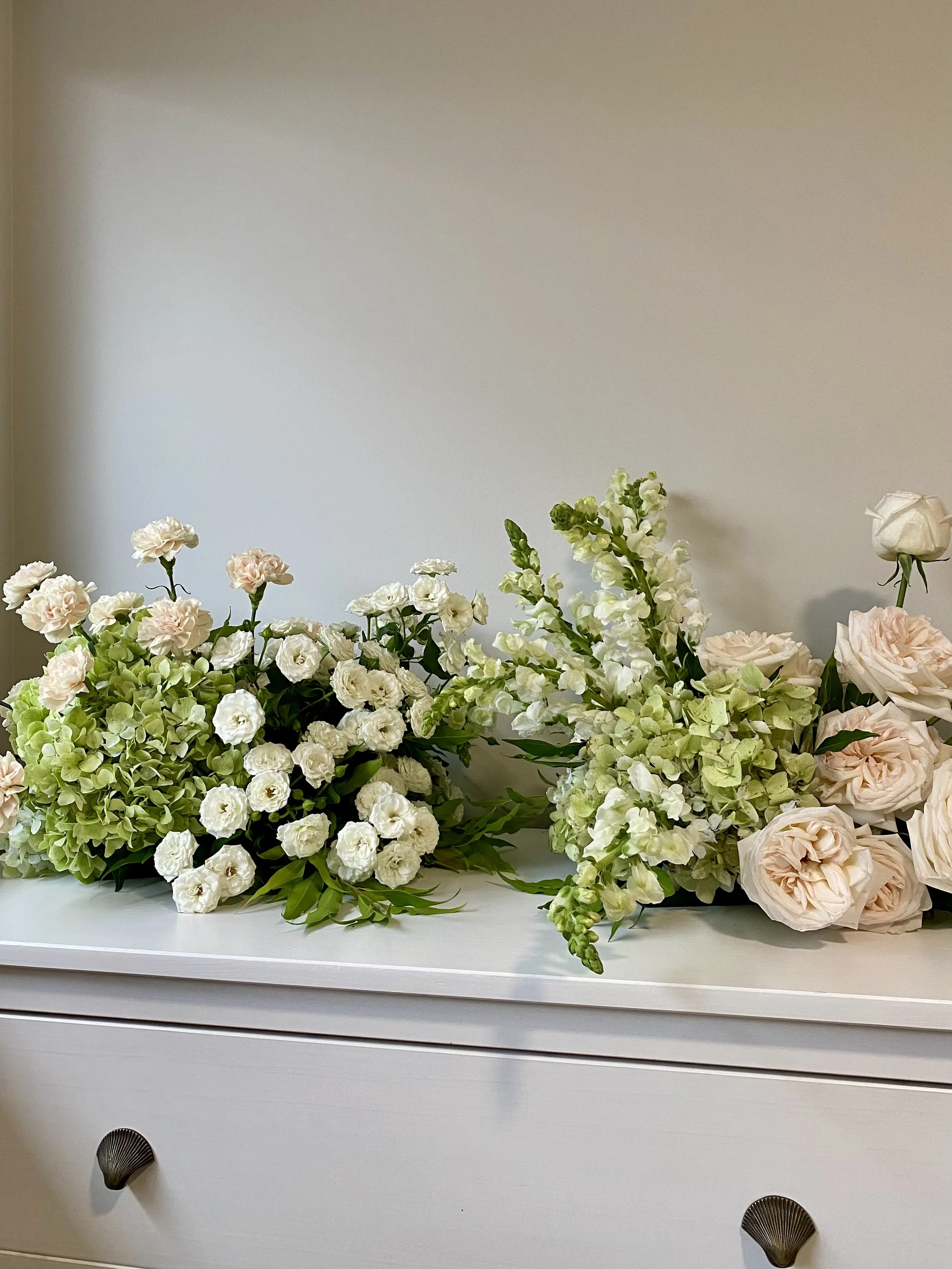 Arrangement of white and pale pink flowers on a white dresser against a plain light-colored wall.