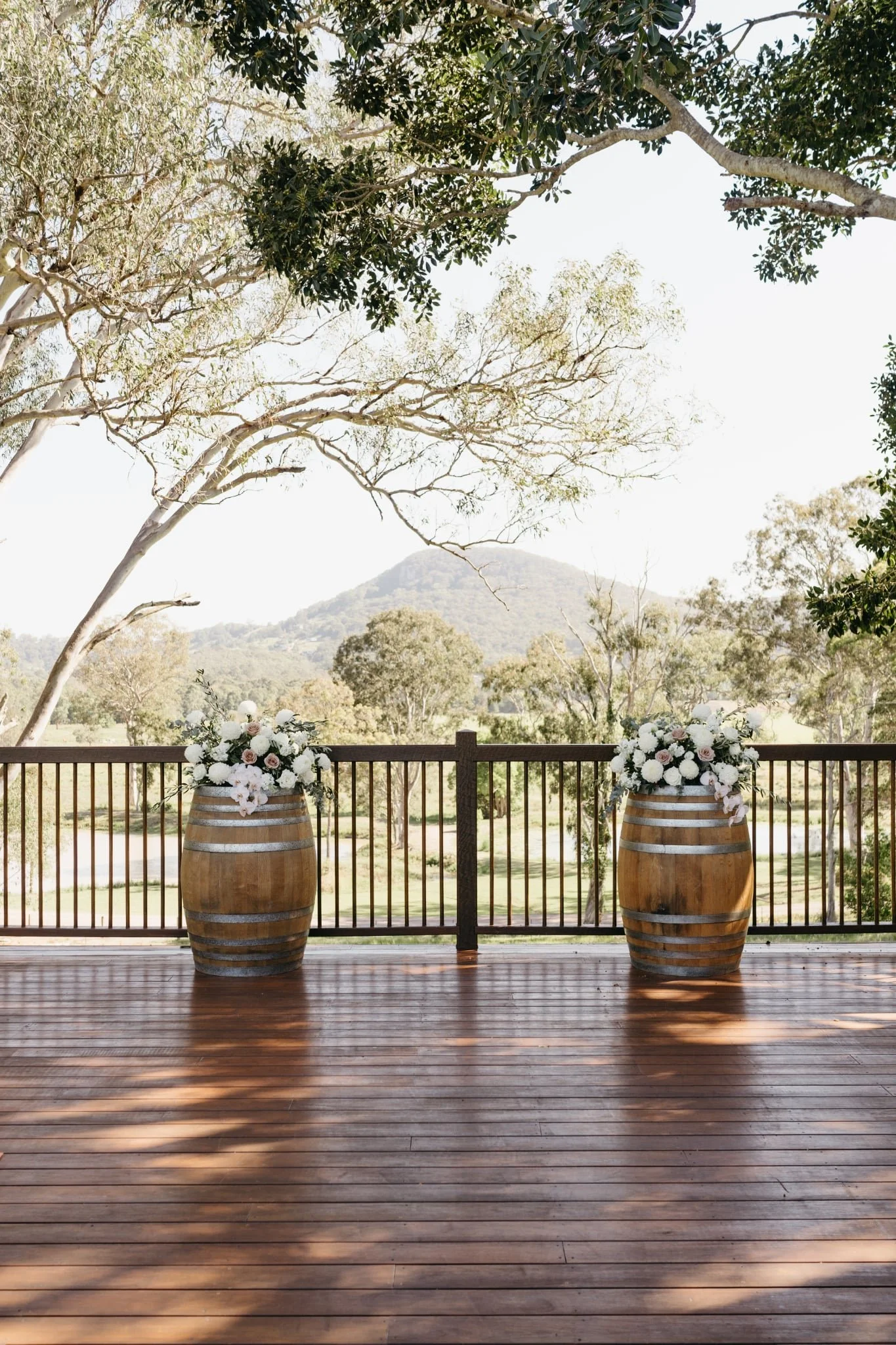Two wooden barrels filled with white and pink flowers sitting on a wooden deck with a scenic view of trees and distant mountains.