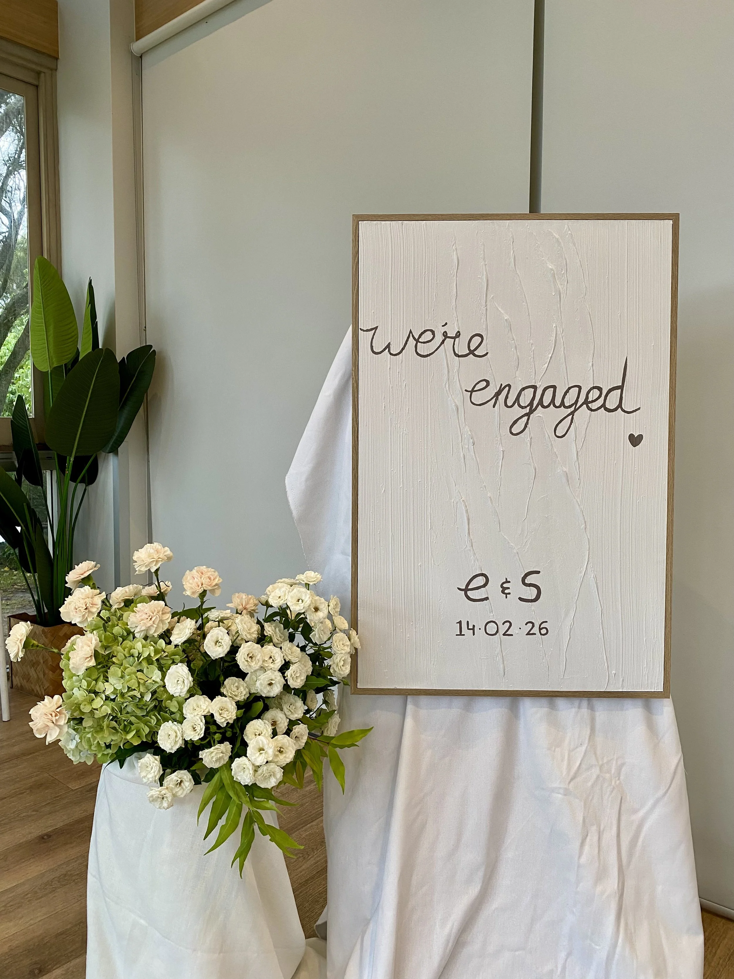 Wedding sign on white backdrop with flower arrangement in a white vase, and a green plant nearby. The sign reads 'we're engaged' with initials 'e & s' and date '14.02.26'.
