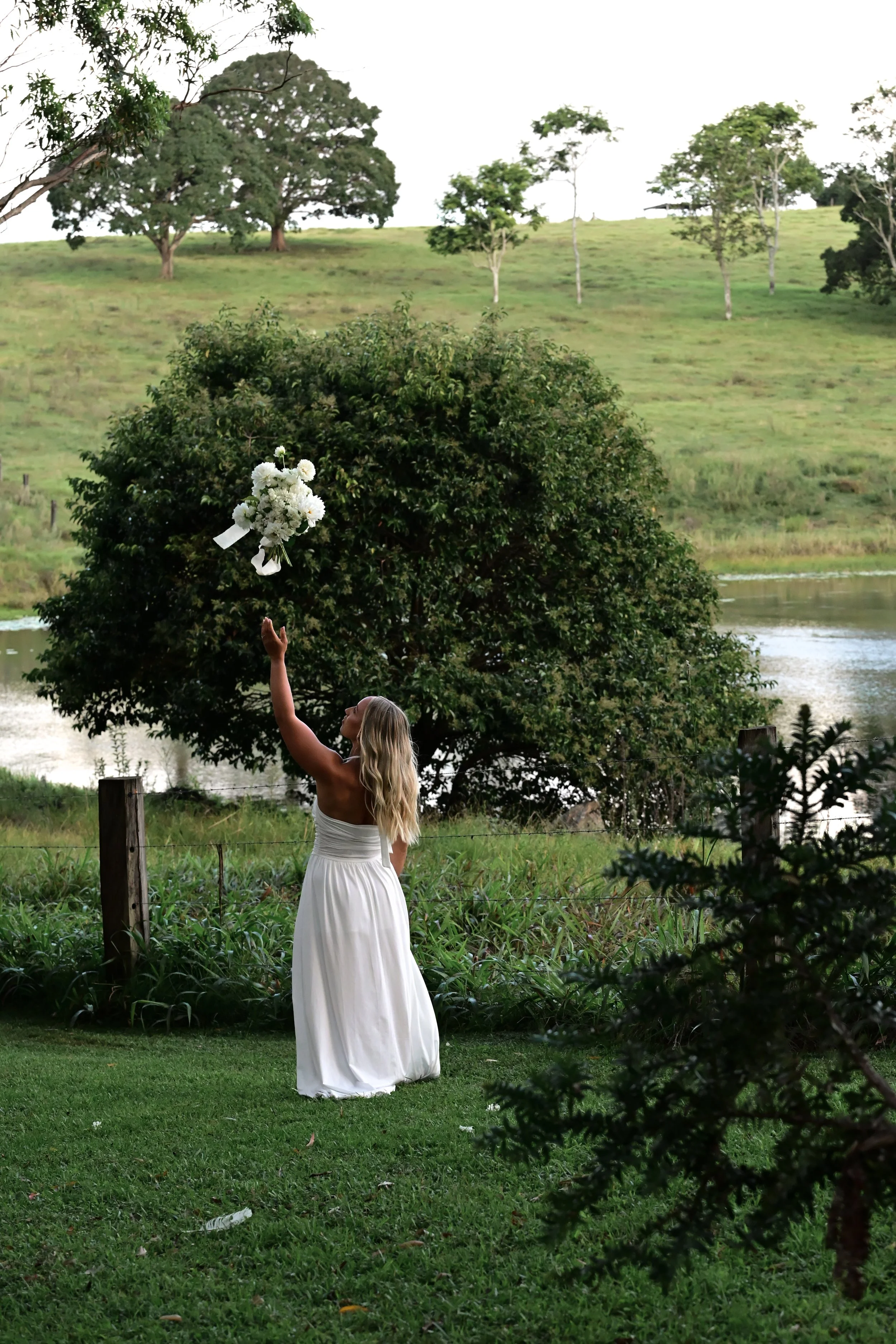 A woman in a white dress stands outdoors near a bush with white flowers, reaching out to throw a bouquet into the air, with a grassy field, trees, and a body of water in the background.