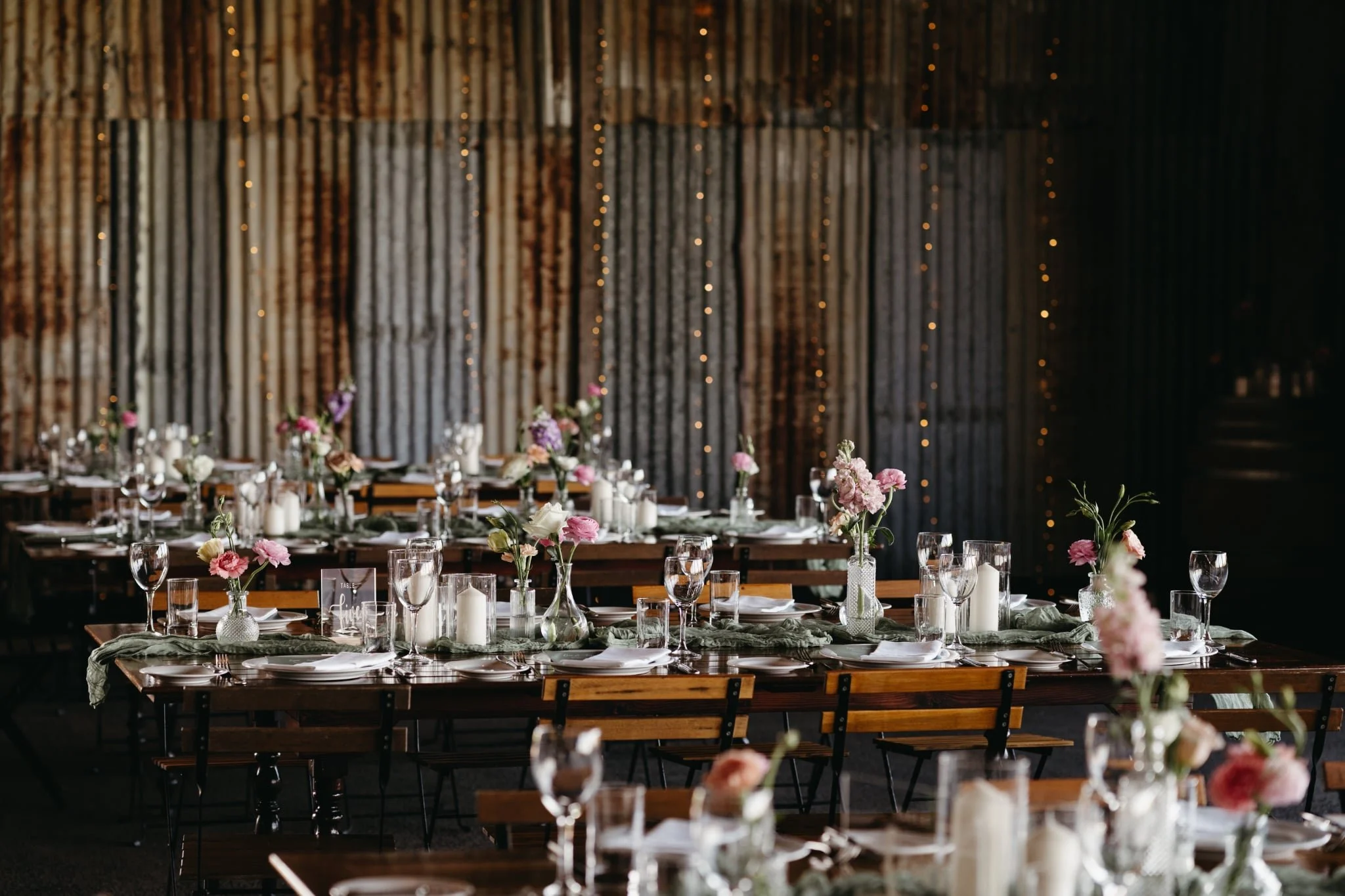 Elegant banquet table setup with floral centerpieces, candles, and glassware, in a rustic venue with string lights and a wooden backdrop.