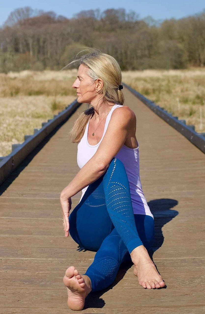 An older woman with gray hair tied back in a ponytail wearing a white tank top and blue leggings practicing yoga outdoors on a wooden bridge with a grassy field and trees in the background.