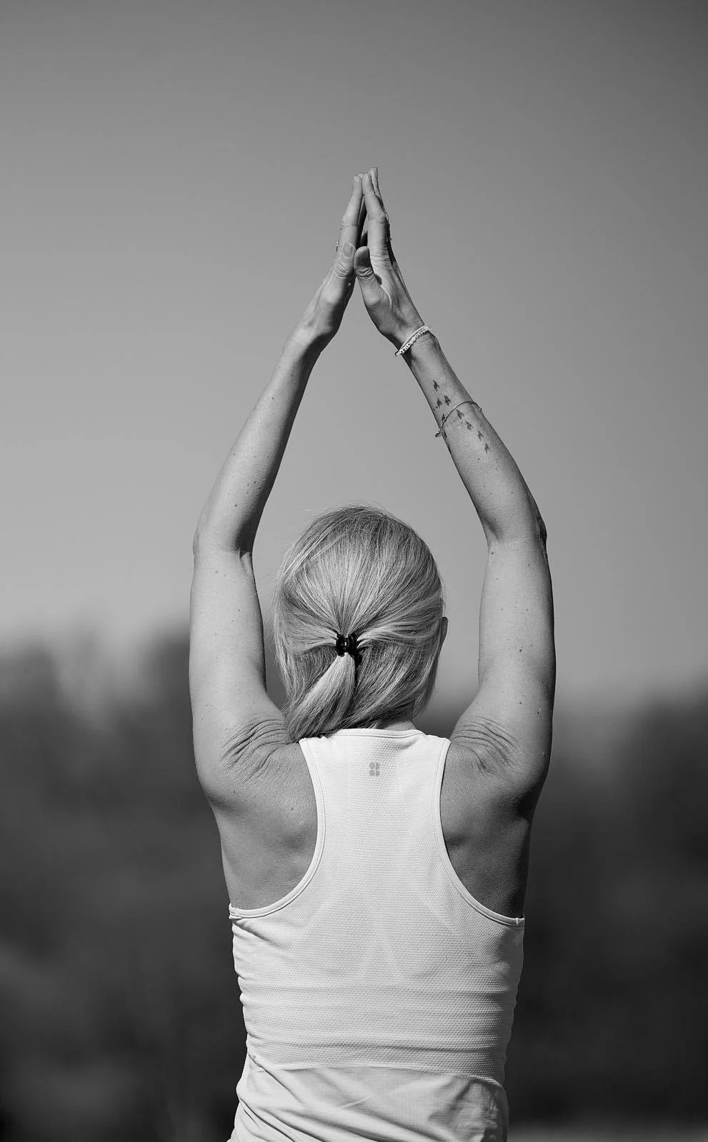 Back of a woman with blonde hair in a ponytail, wearing a sleeveless top, standing outdoors with arms raised above her head in a prayer yoga pose.