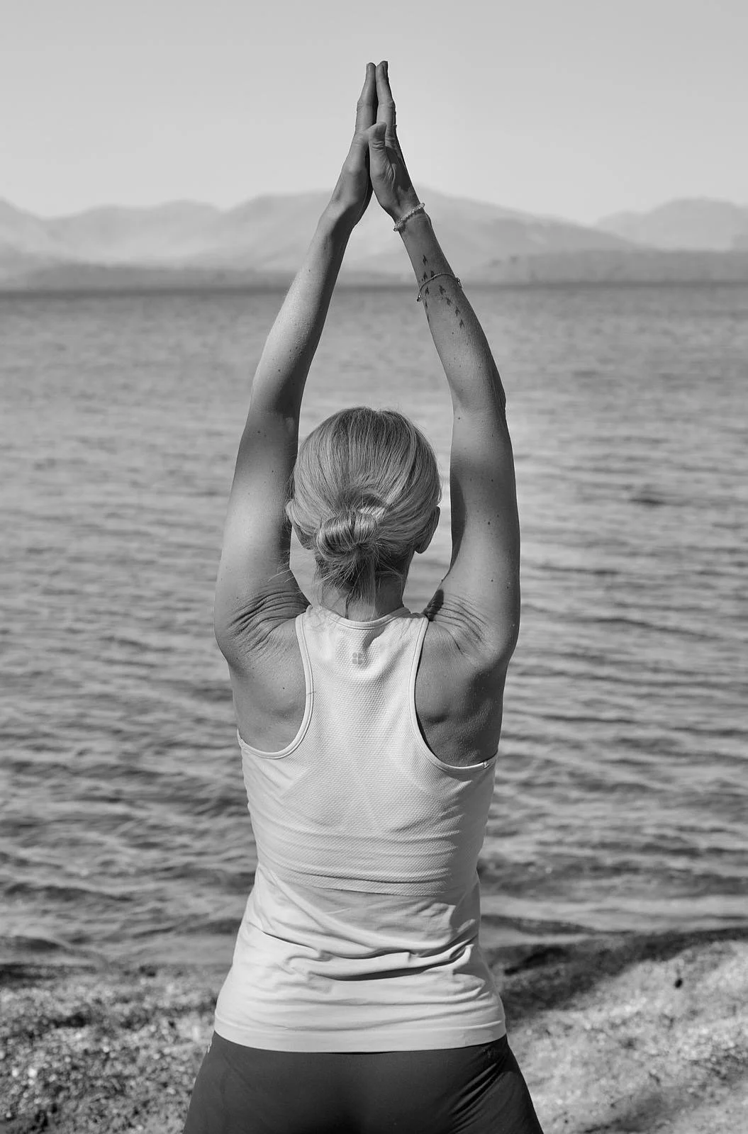 A woman with blonde hair tied in a bun practicing yoga by a body of water, standing with her back to the camera, arms raised overhead in a prayer position.