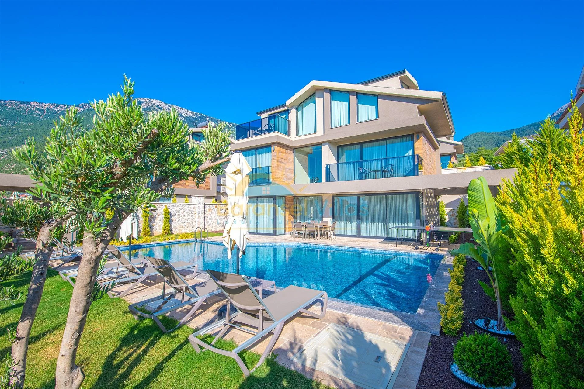 Modern multi-story house with large glass windows, a swimming pool, lounge chairs, and green landscaped yard set against mountain backdrop under clear blue sky.