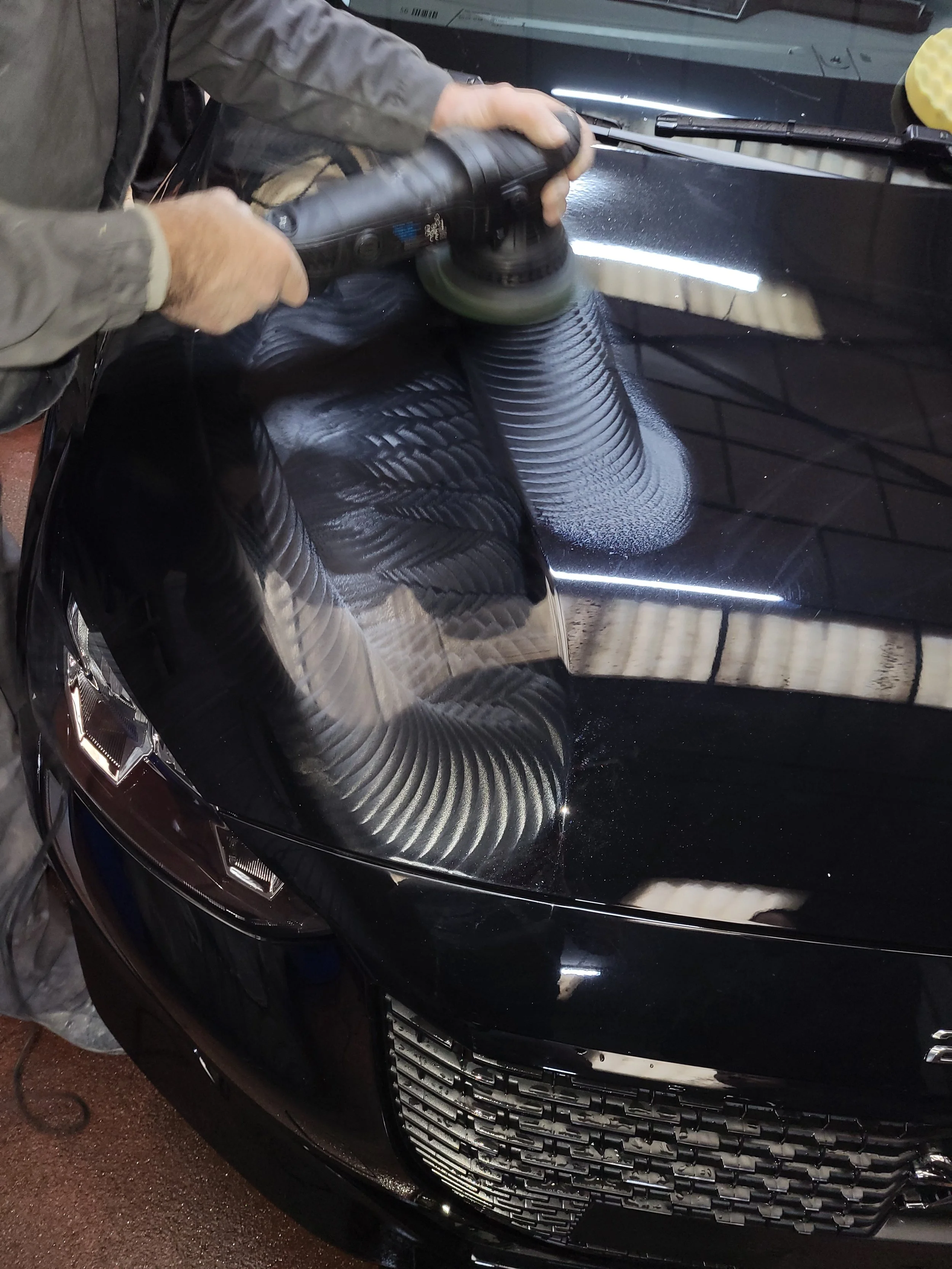 Une personne utilisant une polisseuse sur la surface du capot d'une voiture noire.