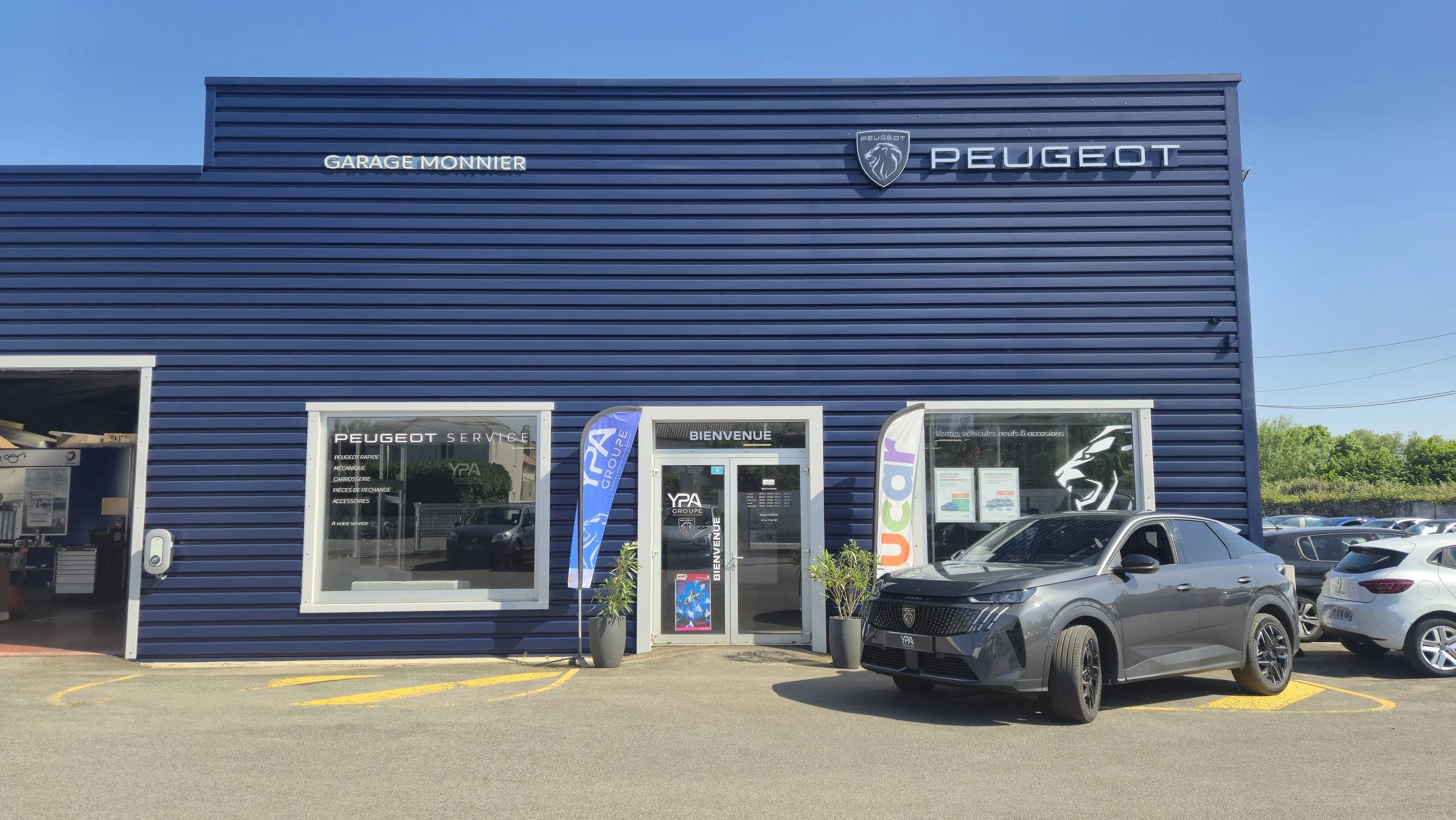 Façade d'un centre de réparation automobile Peugeot avec un bâtiment bleu, un logo Peugeot, une voiture noire garée devant, deux plantes en pots, et plusieurs autres véhicules dans le parking.