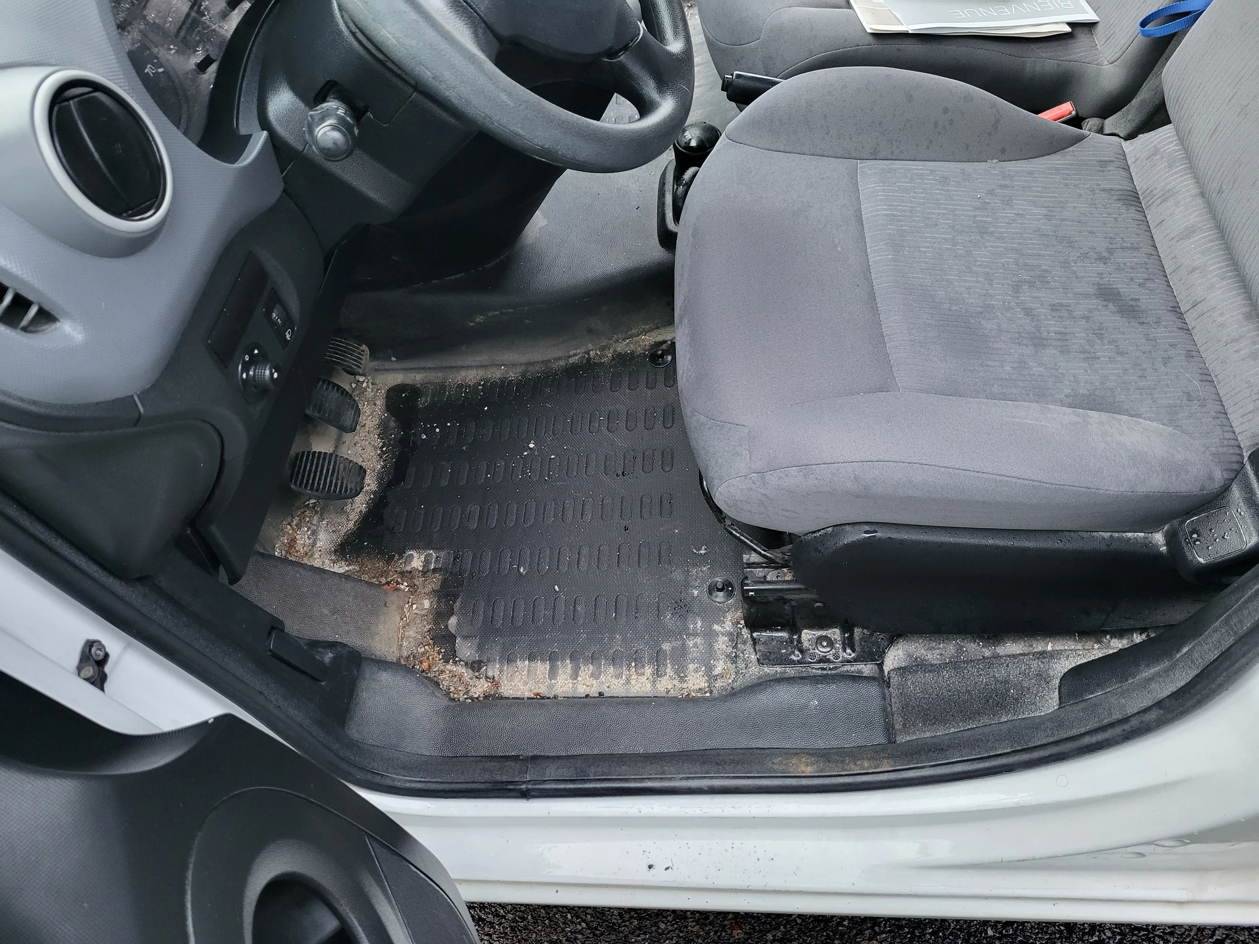 Intérieur d'une voiture montrant le sol sale avec une pédale et un siège en tissu gris.