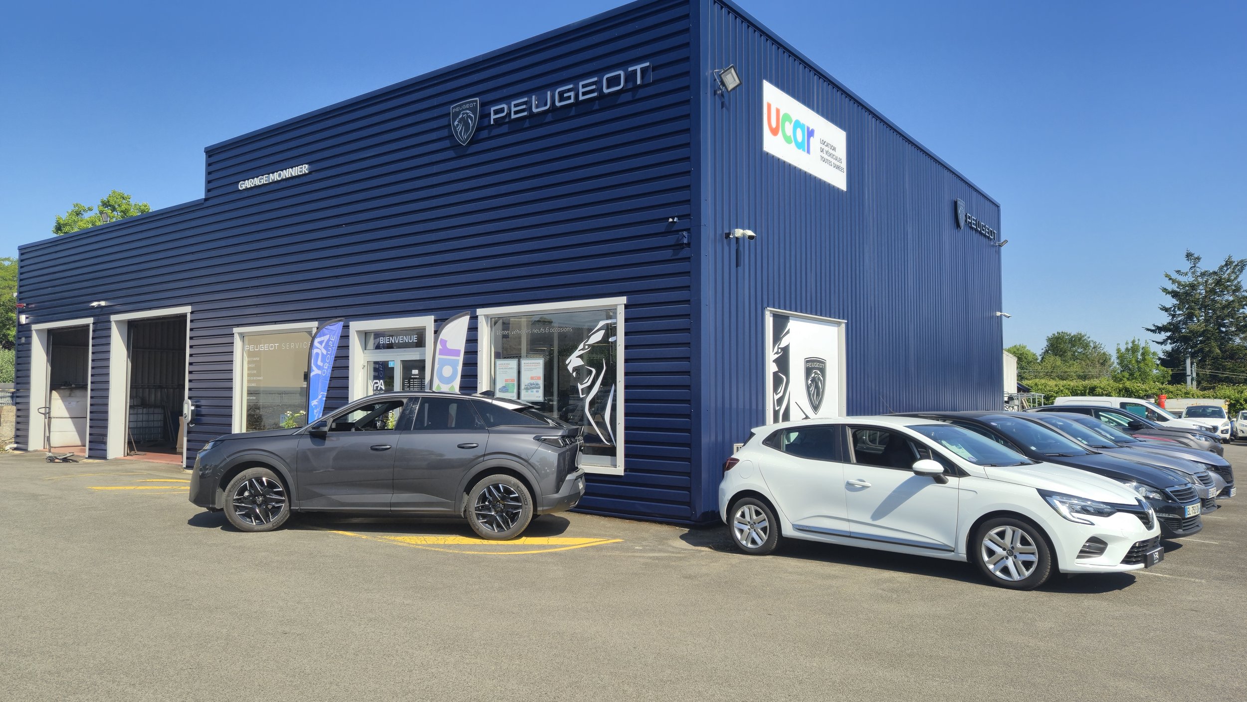 Concessionnaire automobile Peugeot avec plusieurs voitures en extérieur, bâtiment bleu avec logos Peugeot et Ucar, panneaux d'accueil et drapeaux publicitaires.