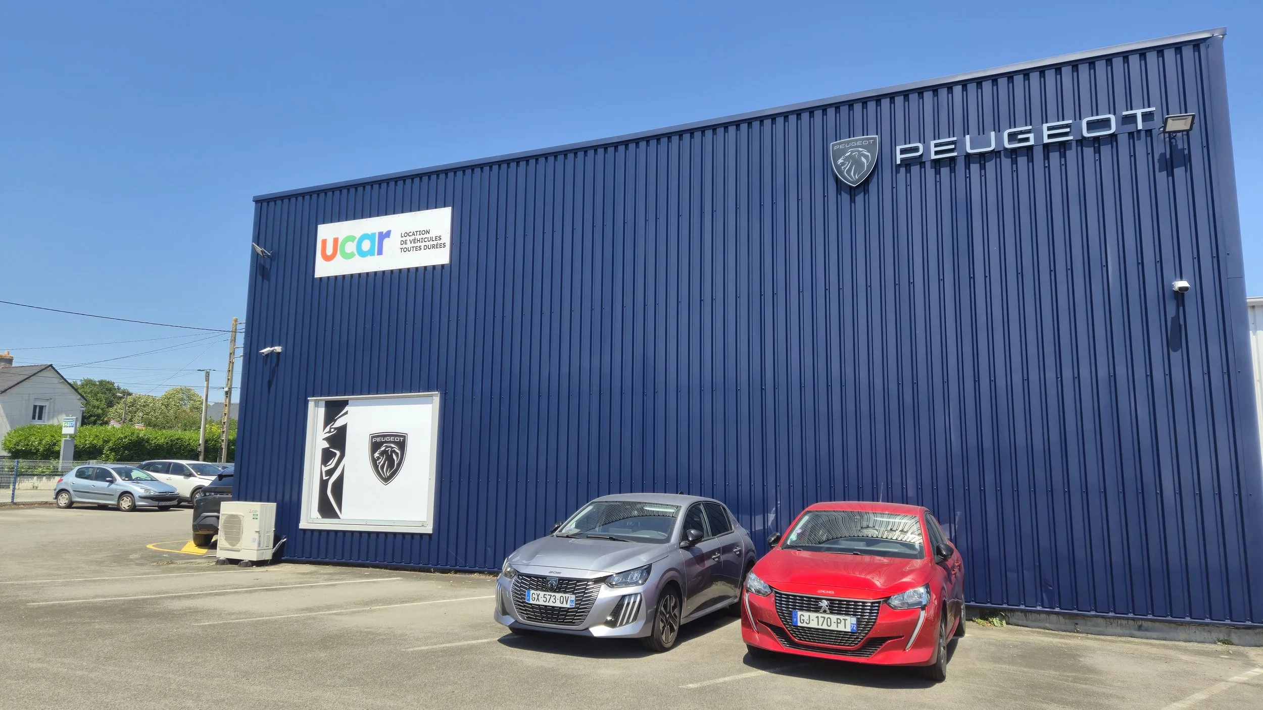 Vue d'une grande façade de bâtiment bleu avec logos Peugeot et UCAR, deux voitures rouges et grises garées devant, ciel bleu clair.