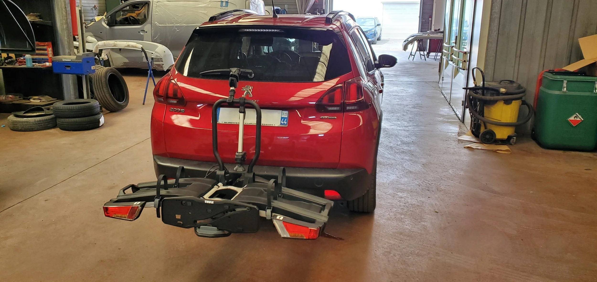 Une photo de l'arrière d'une voiture rouge Peugeot dans un garage, avec un porte-vélos attaché à l'arrière de la voiture.