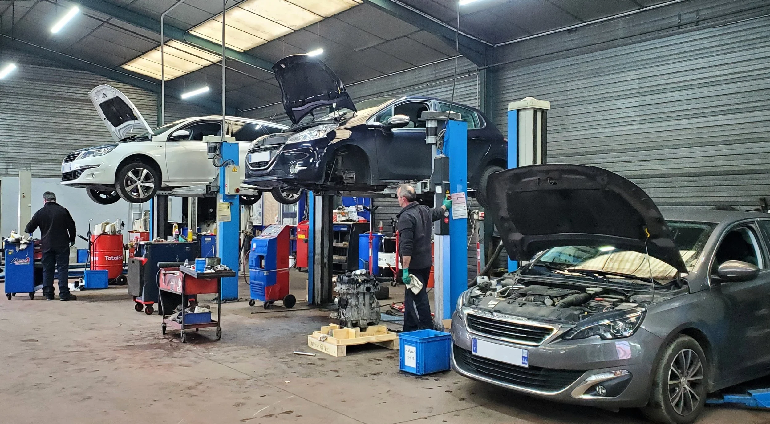 Atelier de réparation de voitures, avec plusieurs voitures en réparation, deux monte-charges accueillant des véhicules, mécaniques en train de travailler sur des véhicules avec capots ouverts.