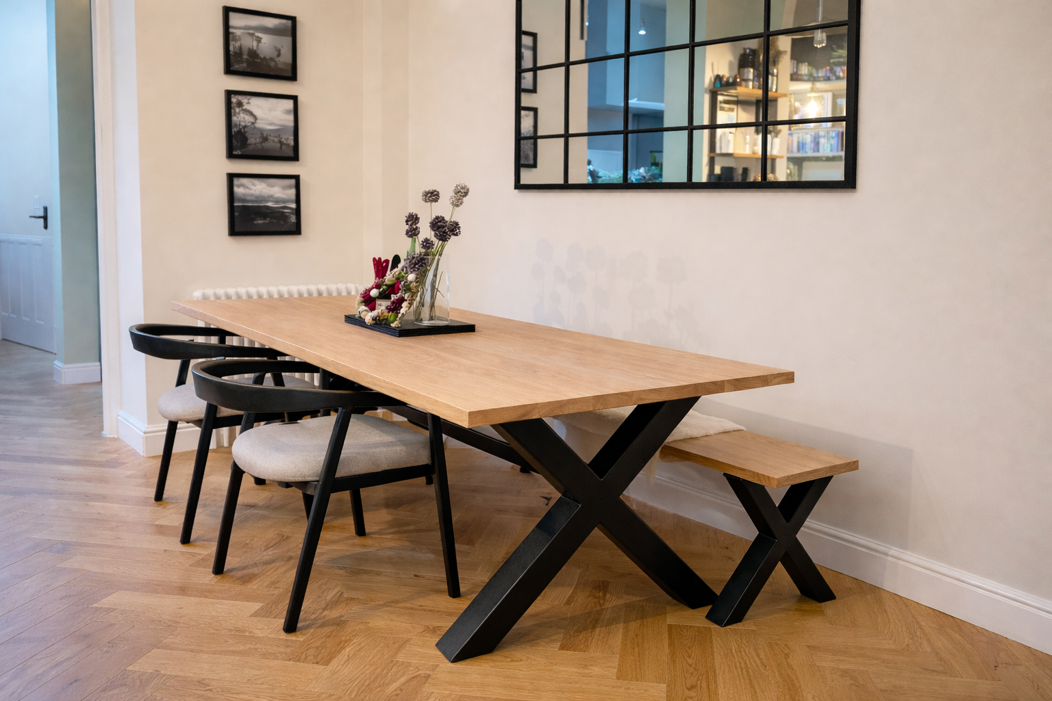 Dining room with a custom wooden dining table, three black chairs with beige cushions, a wooden bench, a vase with flowers, a mirror on the wall, and framed black-and-white photographs.