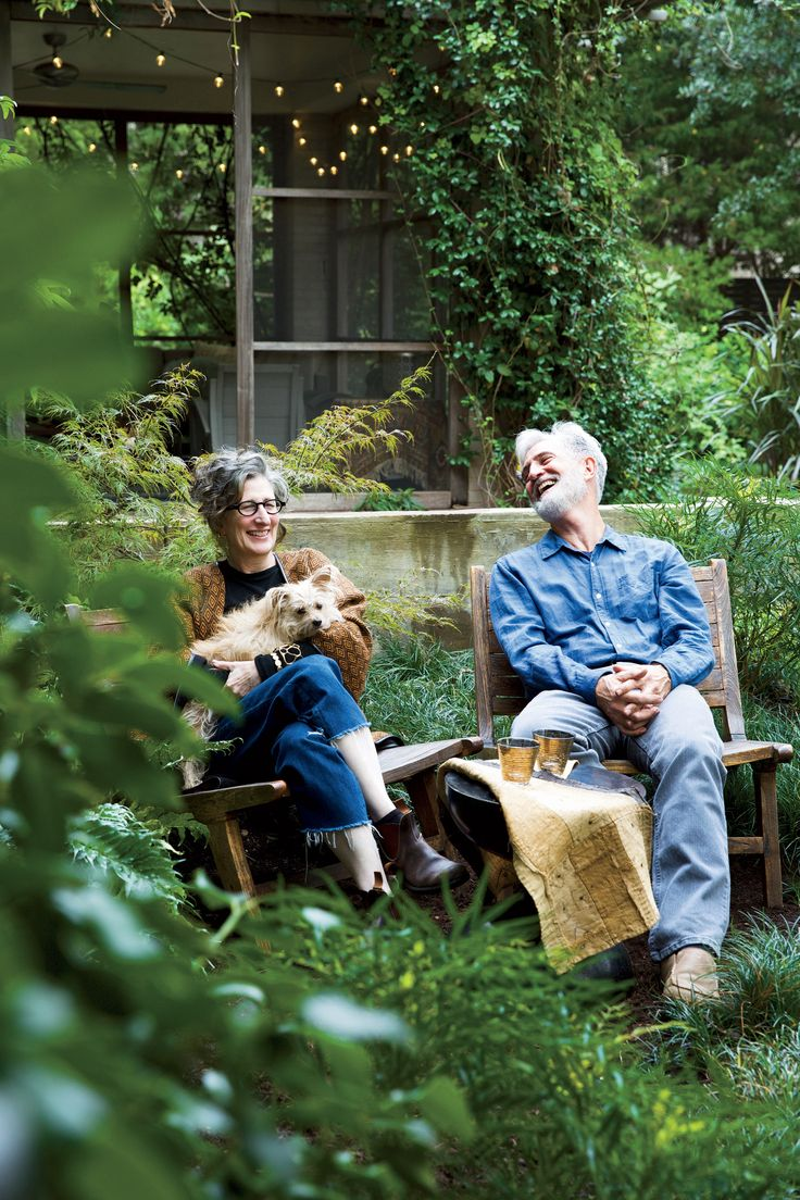 Een moeder en vader zitten ontspannen op tuinstoelen, lachend en pratend, terwijl de moeder hun hondje vasthoudt. Ze zitten in een groene tuin met planten, een houten huis aan de achtergrond en sfeervolle lichtjes.