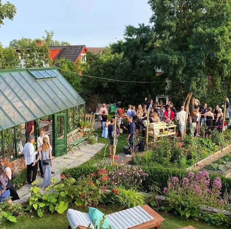 Een tuinfeest met mensen die genieten in een bloeiende tuin met een kas en groene planten.