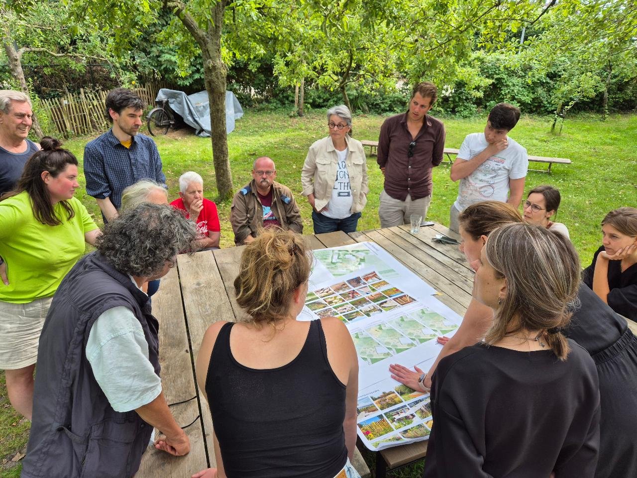 Mensen verzamelen zich rondom een houten tafel in een tuin, waar plannen en afbeeldingen liggen, waarschijnlijk voor een project of ontwerpbespreking.