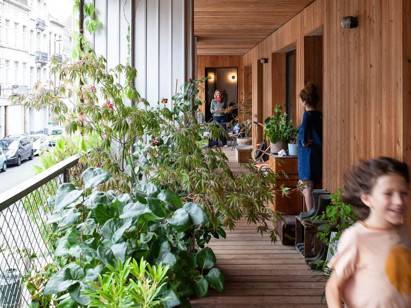 Een overdekt balkon met houten wanden en vloer, versierd met diverse groene planten en slippers, met enkele volwassenen en kinderen die in de buurt zijn.