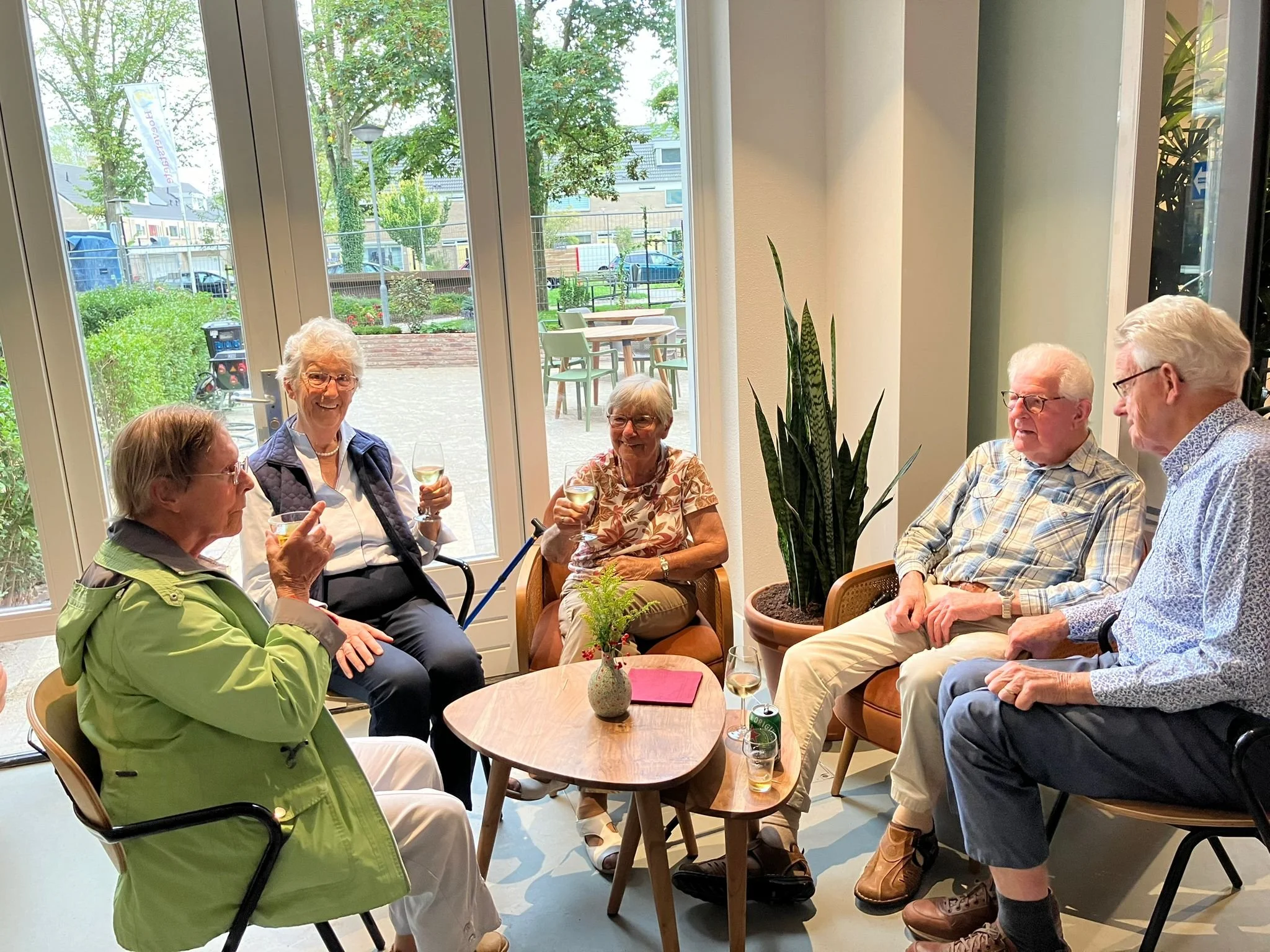 Vijf oudere mensen zitten binnen in een gezellige ruimte, drinken witte wijn, en hebben een gesprek. Ze zitten rond een kleine houten tafel met een plantenpot en een rode map erop. Grote ramen bieden uitzicht op een tuin met bomen en een straat buiten.
