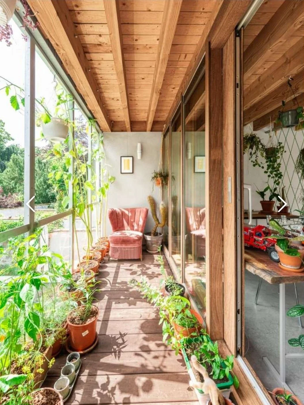 Een gezellige overdekte serre of balkon met veel groene planten, een roze vintage stoel, en houten balkons. Veel natuurlijk licht komt binnen via grote ramen.