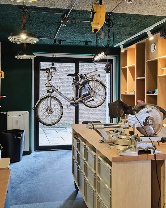 Een werkplaats of fietswinkel met een fiets die aan het plafond hangt, een grote tafel met een snijmachine, en open houten kasten aan de muur. Er is ook een glasdeur naar buiten en wat gereedschap en voorwerpen op de schappen.