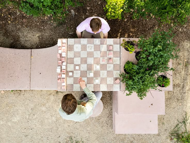 Twee mensen spelen een potje dammen of schaken buiten op een vierkant tafel, met planten en bloemen op een bank ernaast.