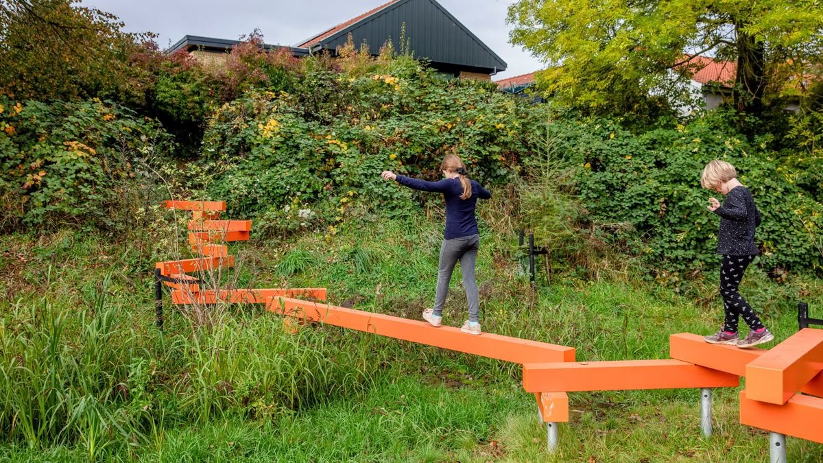 Twee kinderen wandelen over een uitdagende, oranje, wiebelende brug in een groene tuin.