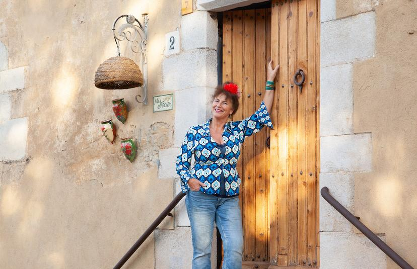 Vrolijke vrouw met een blauwe bloemenblouse en een rode bloem in haar haar, staat buiten bij een houten deur en lacht.