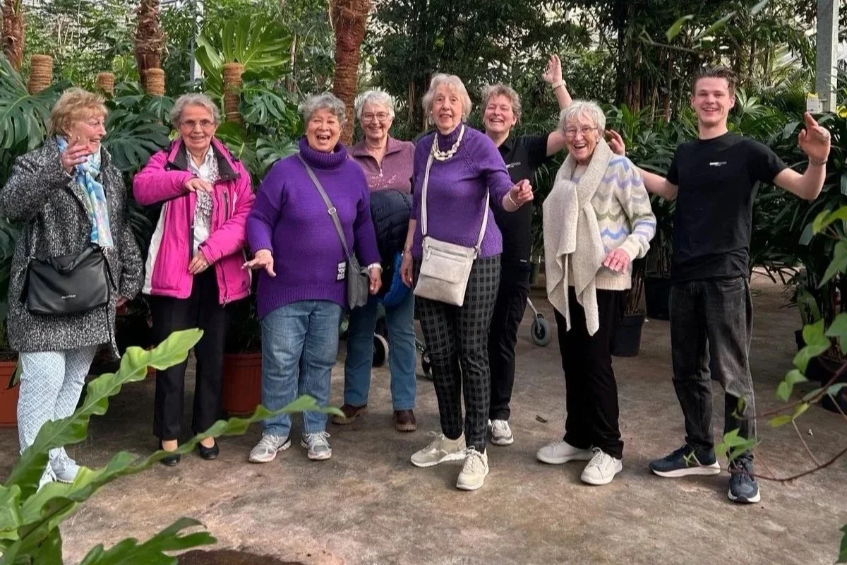 Een groep mensen die in een serre of botanische tuin staan, omringd door grote groene planten, en vrolijk lachen en poseren.