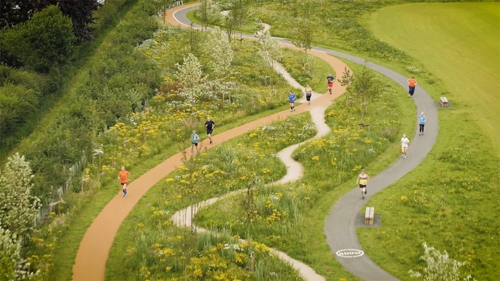 Mensen die joggen en wandelen op een kronkelend pad door een groen park met bloemen en bomen.