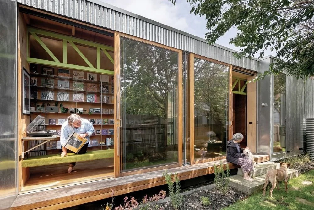 Een modern huis met grote glazen schuifdeuren, een vrouw leest een boek binnen, een oudere vrouw met een hond zit buiten op de stoep en er liggen twee honden in de tuin.