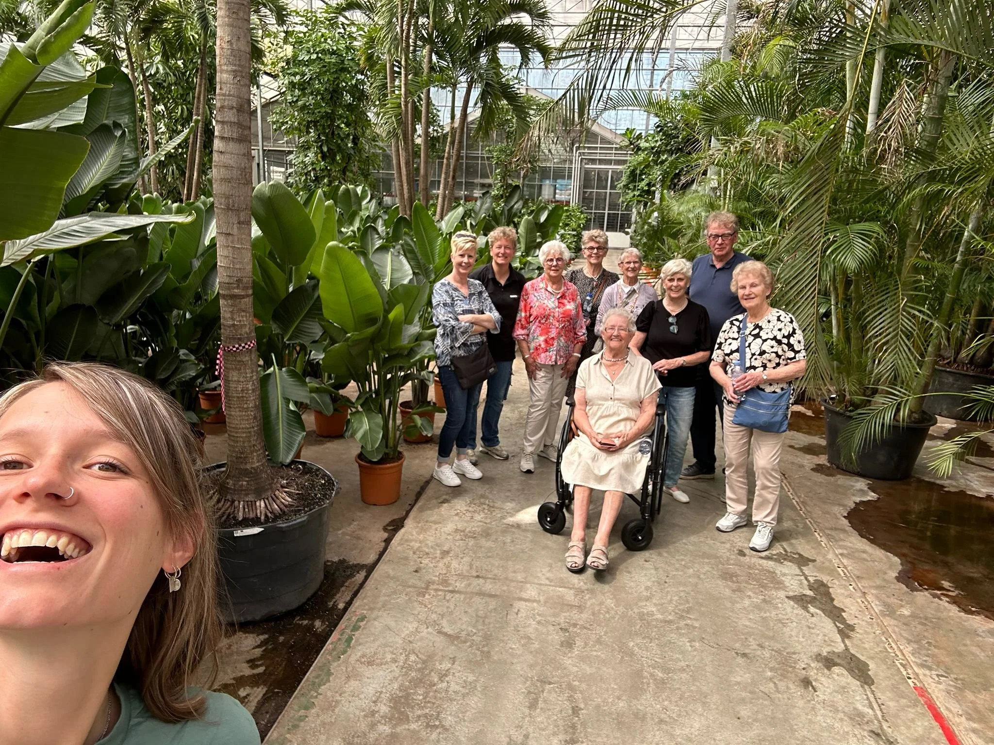 Groep mensen poseert in een tropische kas met grote groene planten, waaronder palmbomen en bananenplanten. Eén vrouw zit in een rolstoel, meerdere mensen staan eromheen, en een jonge vrouw neemt zelf de foto.